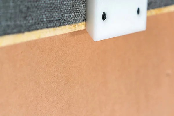 Close view of the back edge of a board showing the fabric wrapped over the solid wood frame, the hardboard back panel, and one white cleat block fixed with two screws