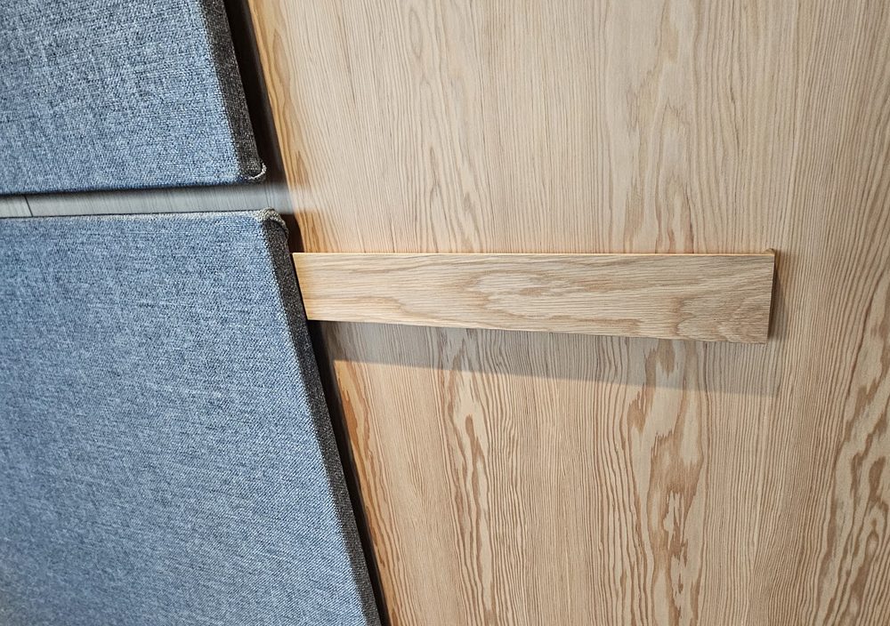 Close view of an oak mounting rail fixed to a panelled wall, with the wrapped edges of two fabric boards meeting it on the left