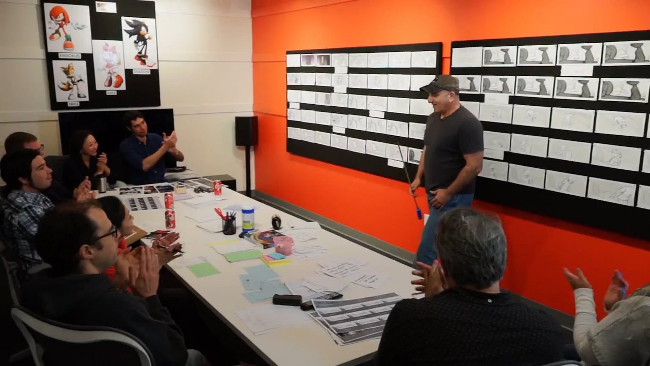 A story review in a studio: a team seated at a long table facing two large black CinemaBoards covered with rows of printed storyboard panels, with one person standing at the wall holding a pointer